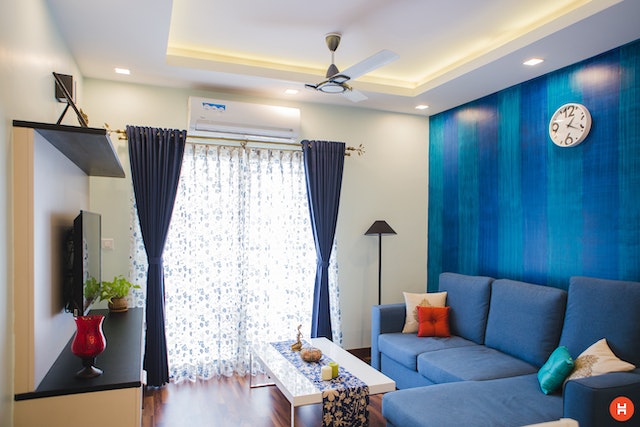 a living room with a blue accent wall in a thousand oaks rental property there is a blue sectional and a square white coffee table facing a television natural light streams in through the white curtains and lights up the space